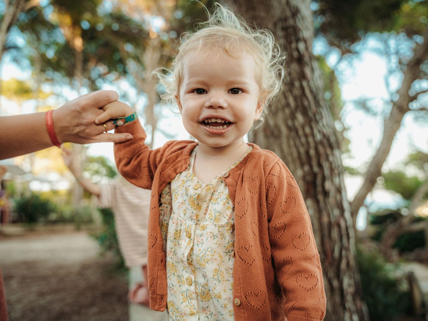 Cheerful blonde toddler girl smiling while adult hand offers something outdoors among pine trees.