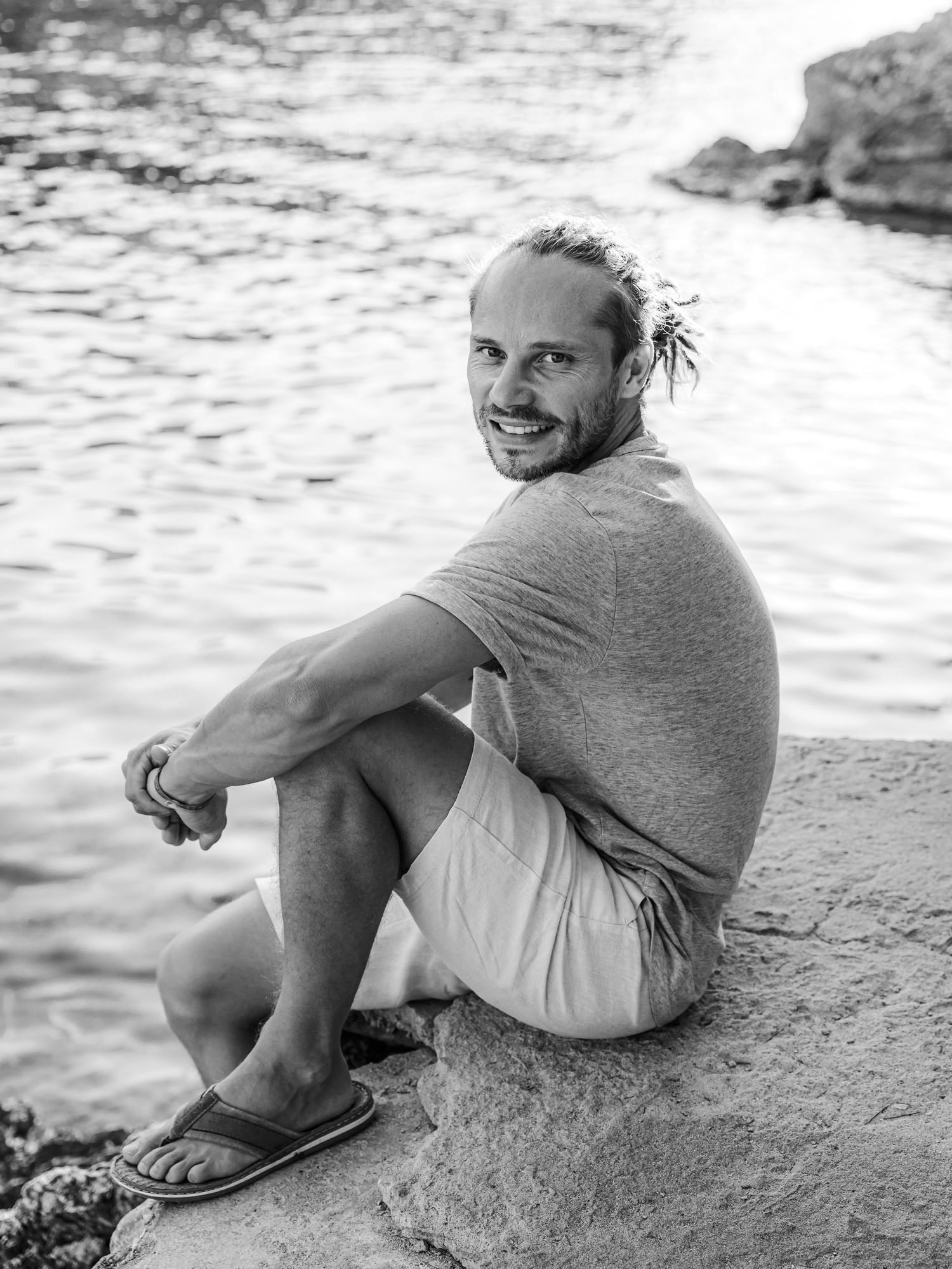 Black and white portrait of a smiling man with a bun sitting on rocks beside a glistening body of water.