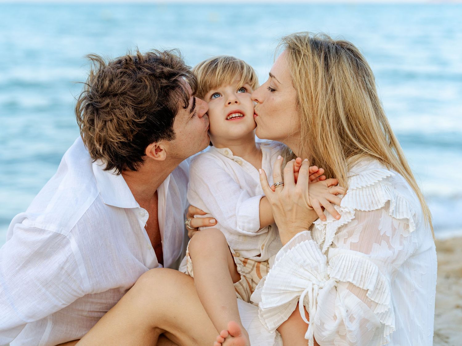 Family beach portrait with parents kissing toddler on cheeks while sitting on sandy shore near ocean.