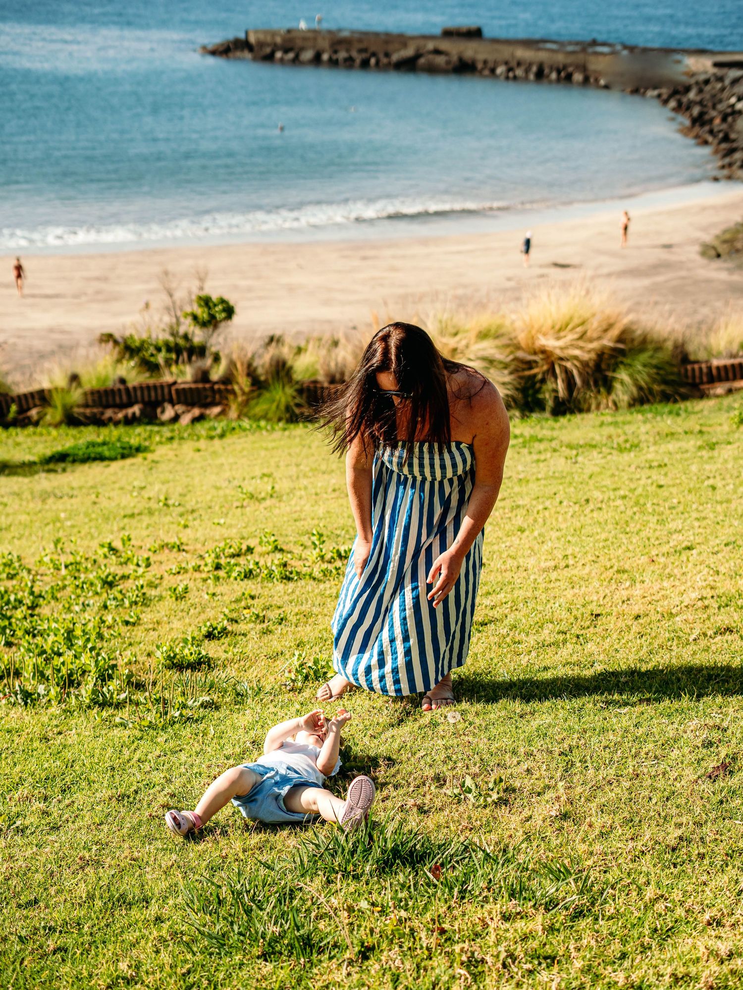 Mother in striped dress helping her toddler on a grassy hillside overlooking a sunny beach and ocean.