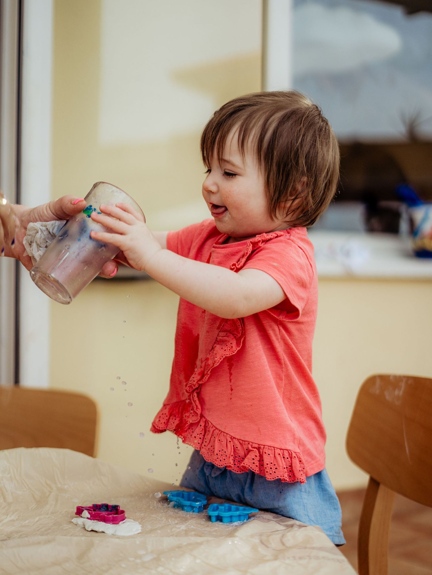 Toddler girl in red top pouring sand from a clear cup onto a table indoors.