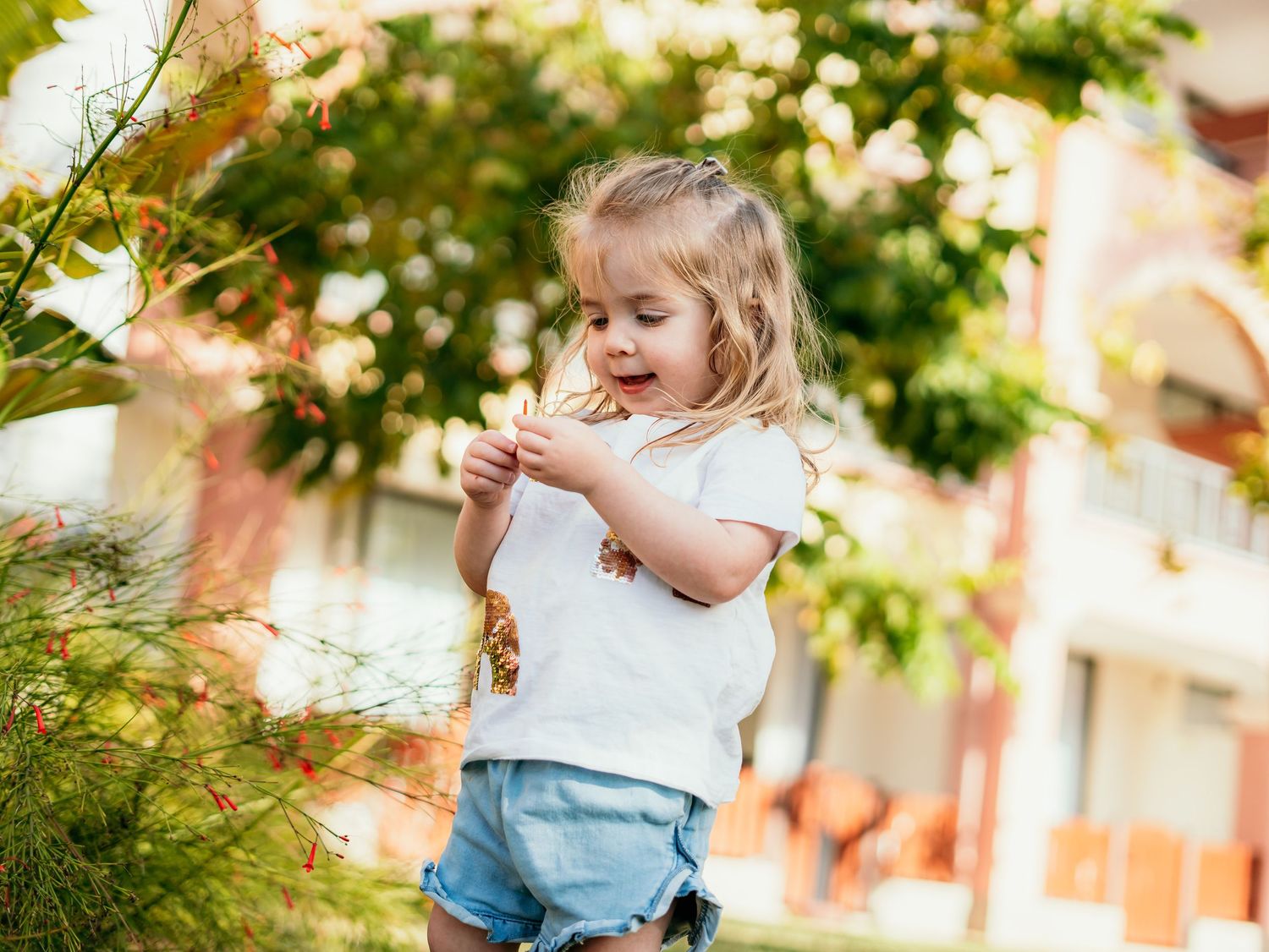 Young toddler girl in white tee and denim shorts playing near green trees and shrubs outdoors.