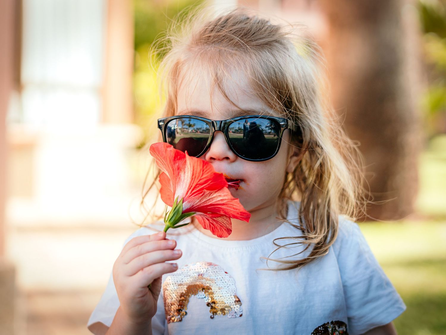Adorable toddler in oversized sunglasses smells a bright red hibiscus flower outdoors.