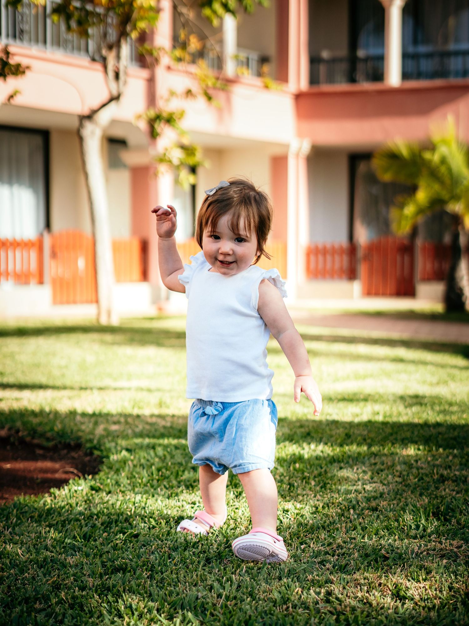 Cheerful toddler girl in white top and blue shorts walking on green grass outside.