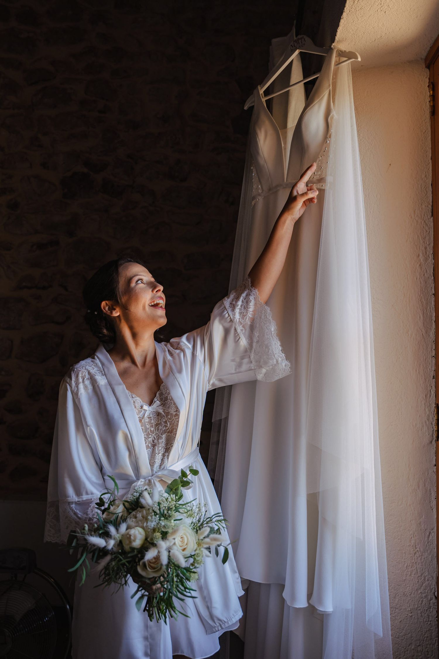 Bride in white robe gazing up at wedding dress hanging on stone wall.