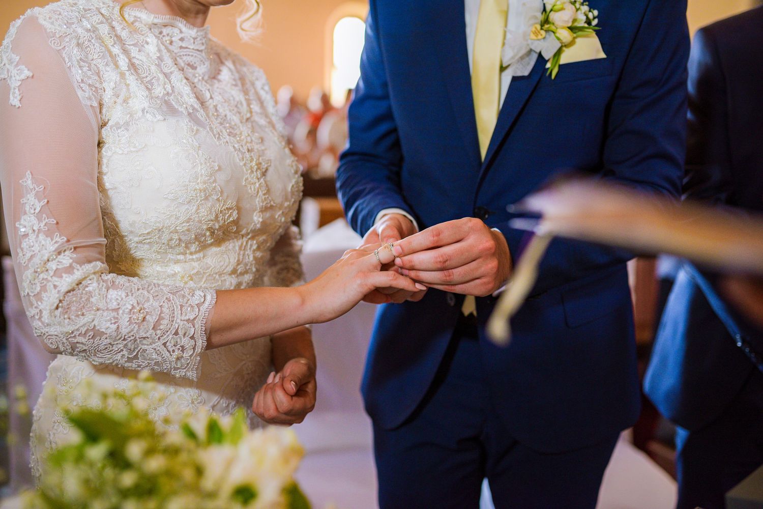 Close-up of bride placing gold wedding ring on groom's finger during indoor ceremony.