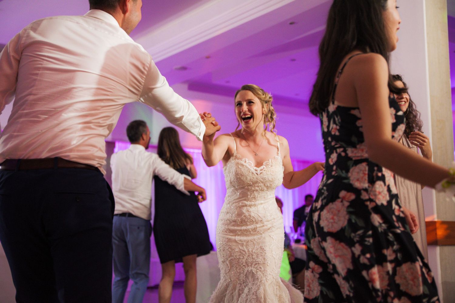 Laughing bride in lace gown dances with guests under purple lights at wedding reception.