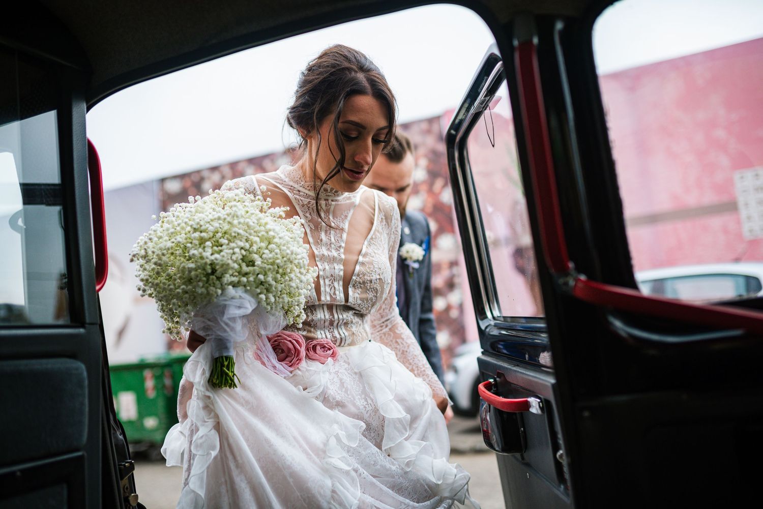 Bride in lace wedding gown stepping out of a vintage black taxi cab holding a baby's breath bouquet.