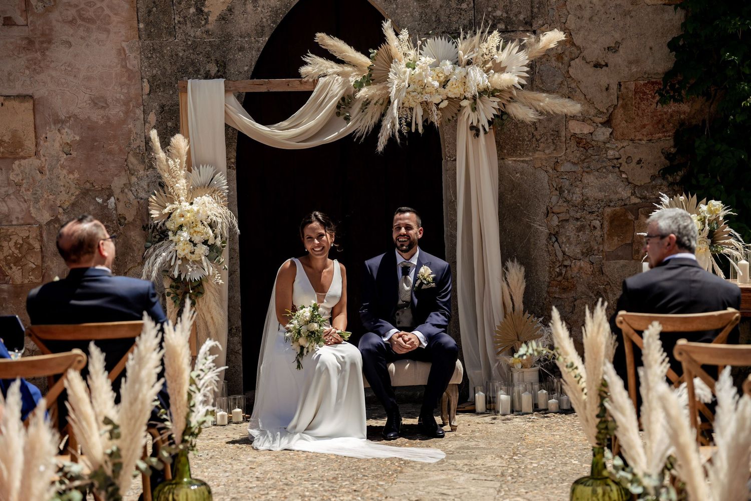 Bride and groom seated at outdoor ceremony beneath elegant pampas grass and dried floral arch.