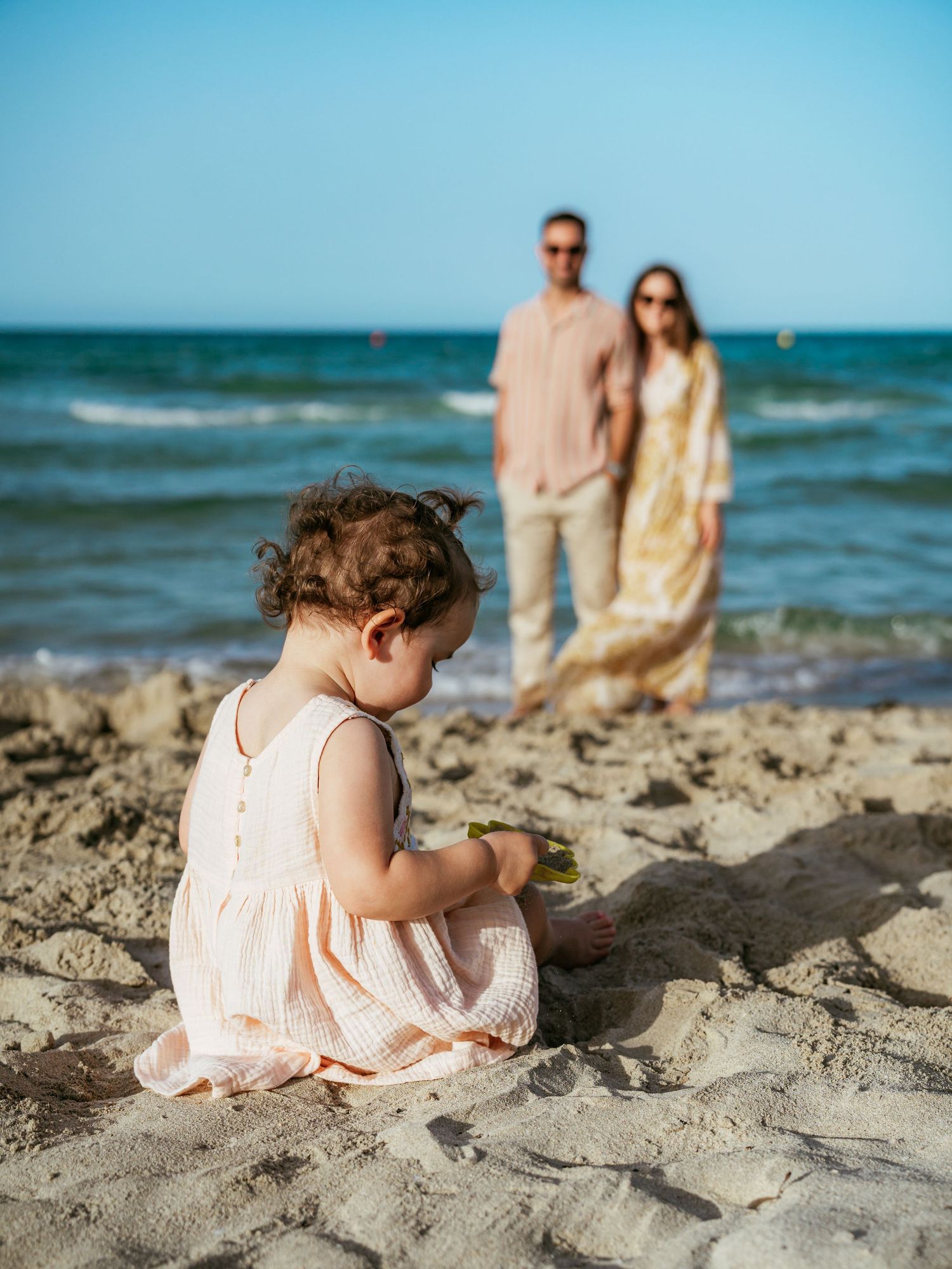Toddler girl in pink dress plays in beach sand while her parents stand together by the ocean behind her.