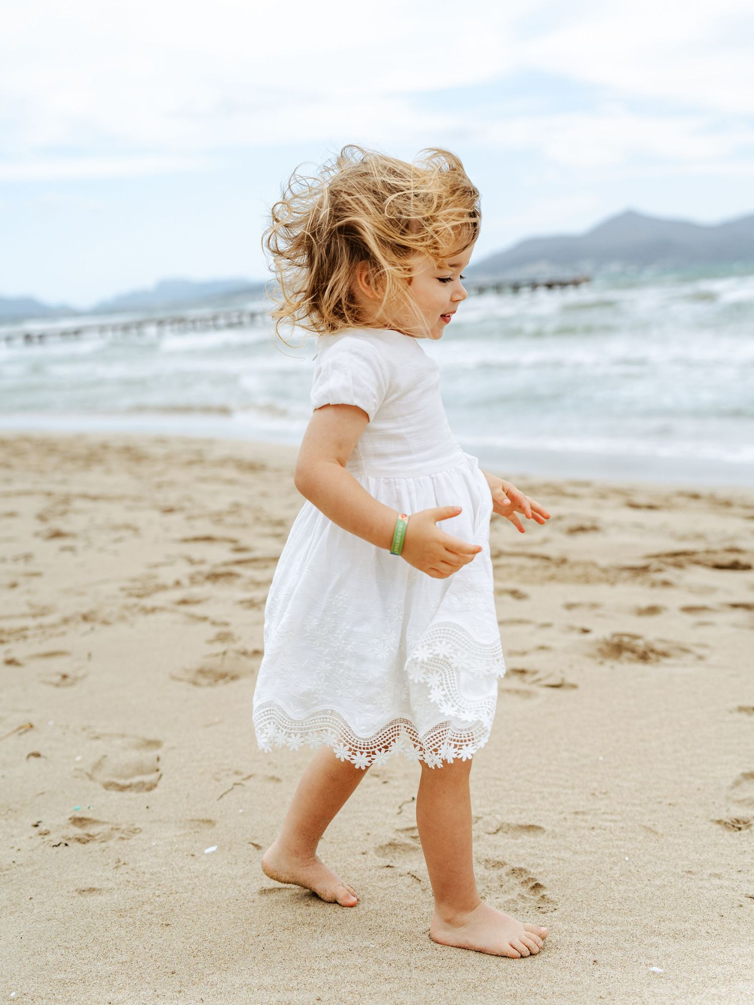 A curly-haired toddler girl in a white lace dress walks barefoot on a sandy beach near the ocean.