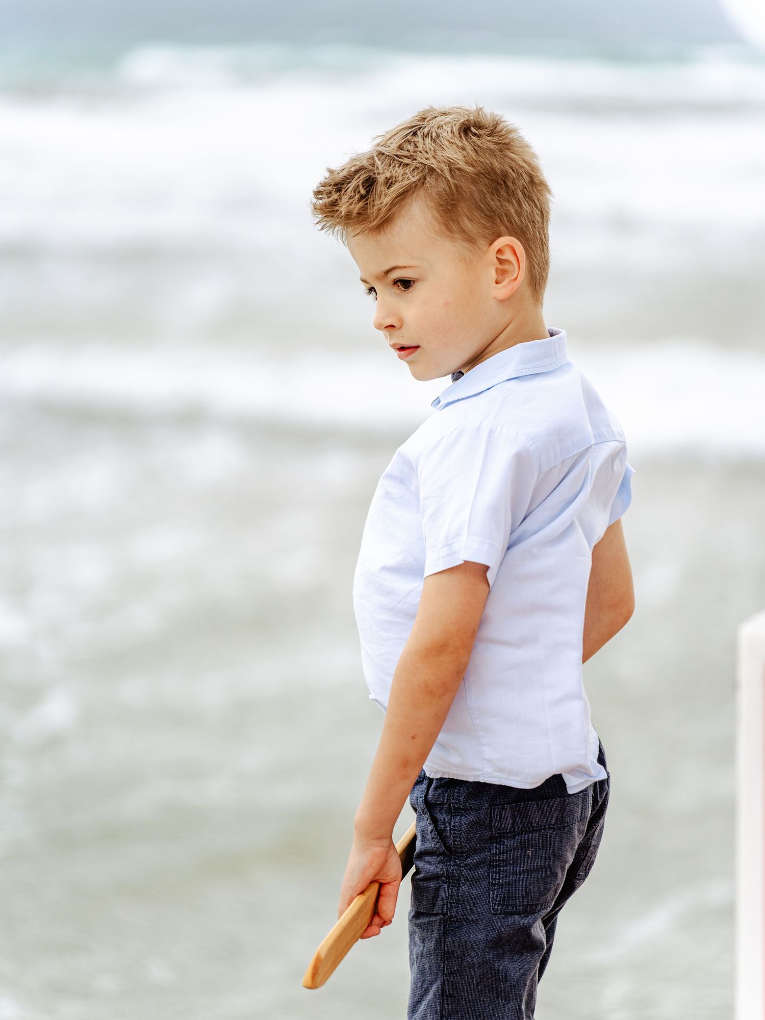 Young blond boy in white shirt holding wooden stick on windy beach.