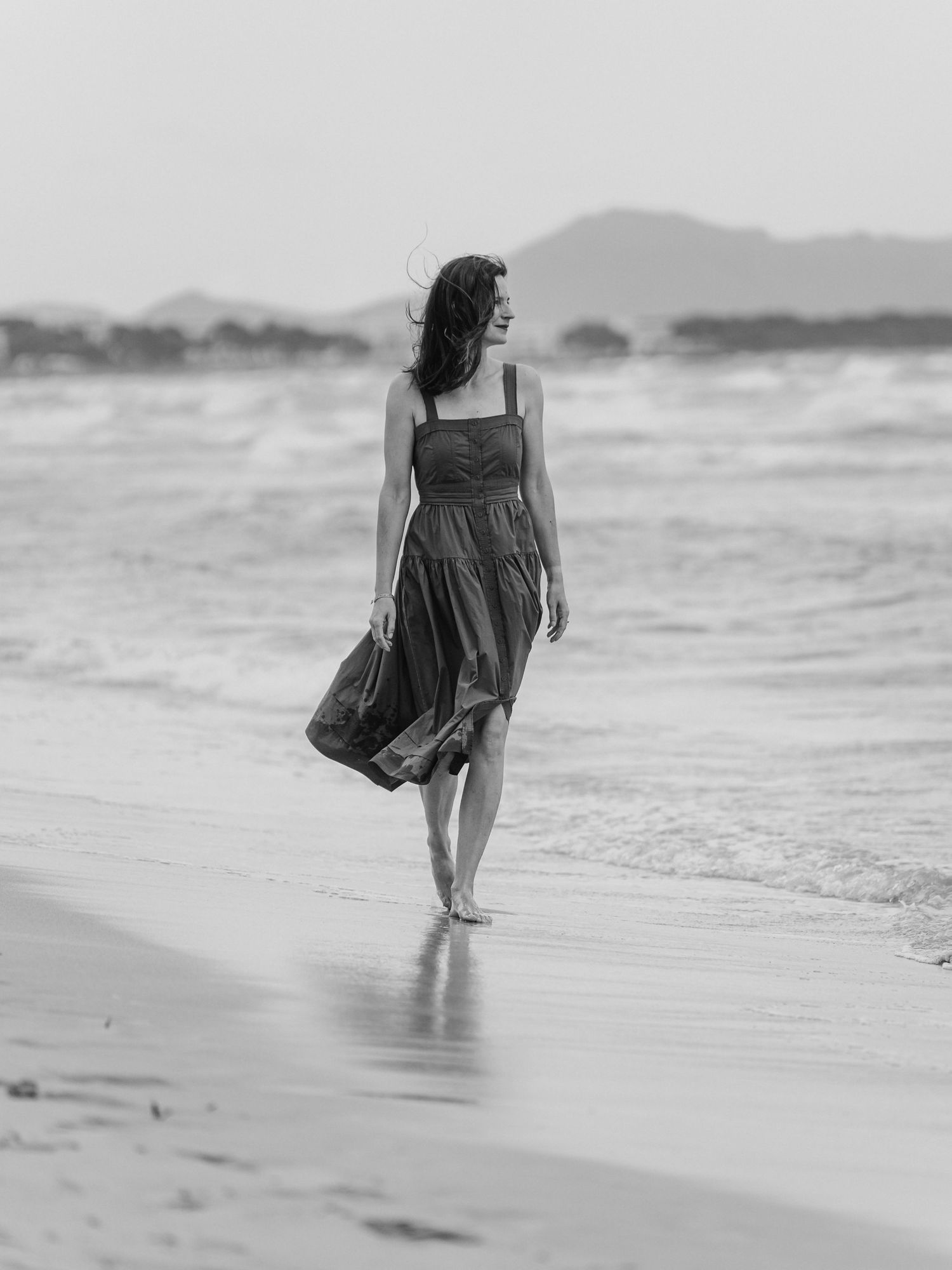 Woman in a dress walking barefoot along a beach in black and white.