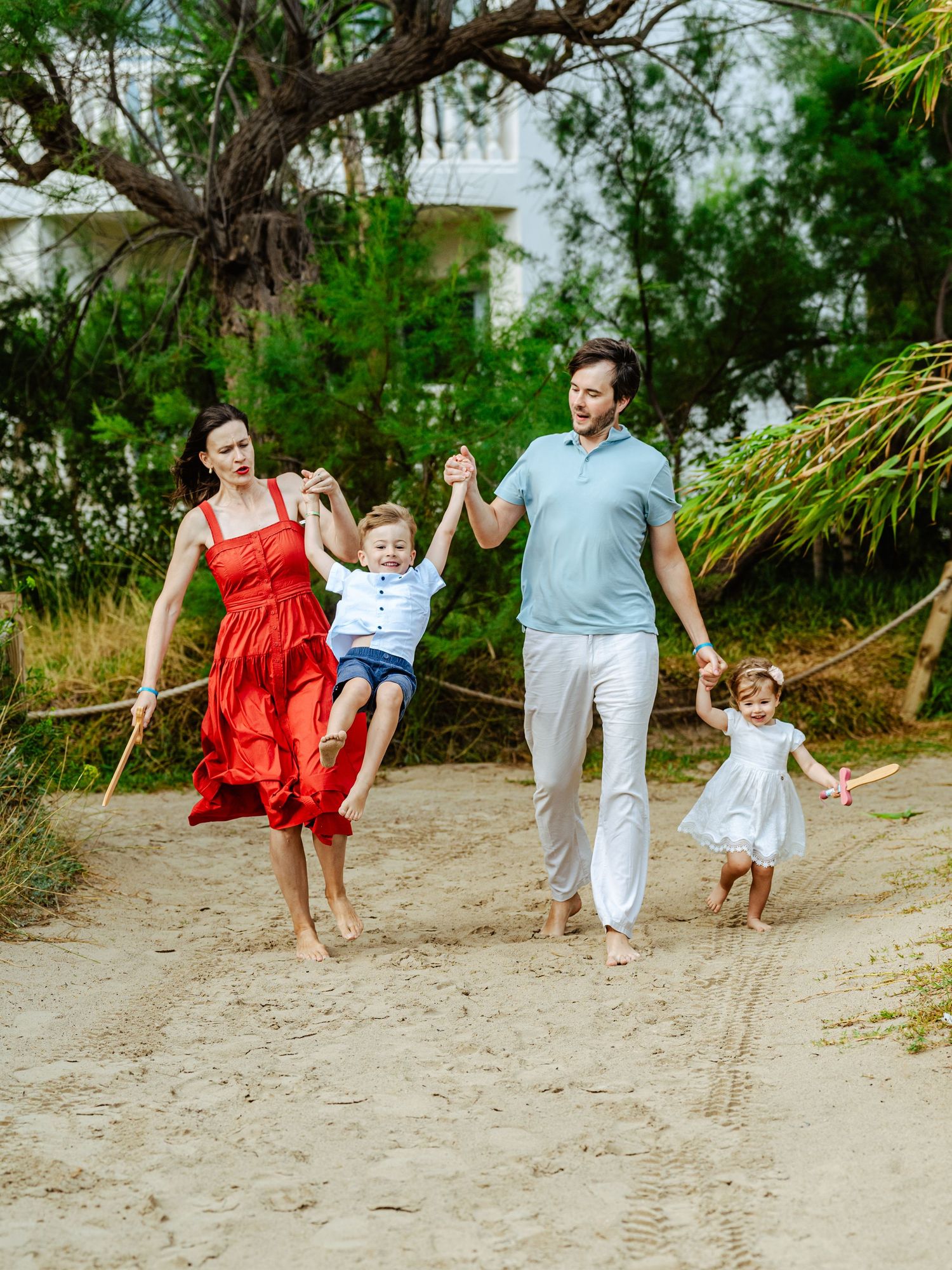 Joyful family of four playing on sandy beach path, mother in red dress swinging boy in the air.