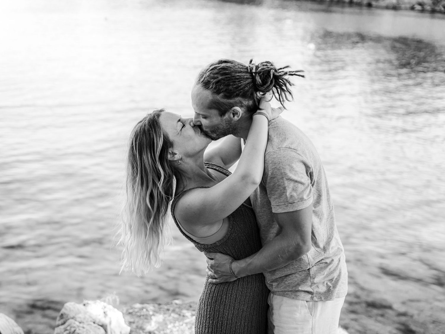 Black and white photo of a couple kissing romantically by the water's edge.
