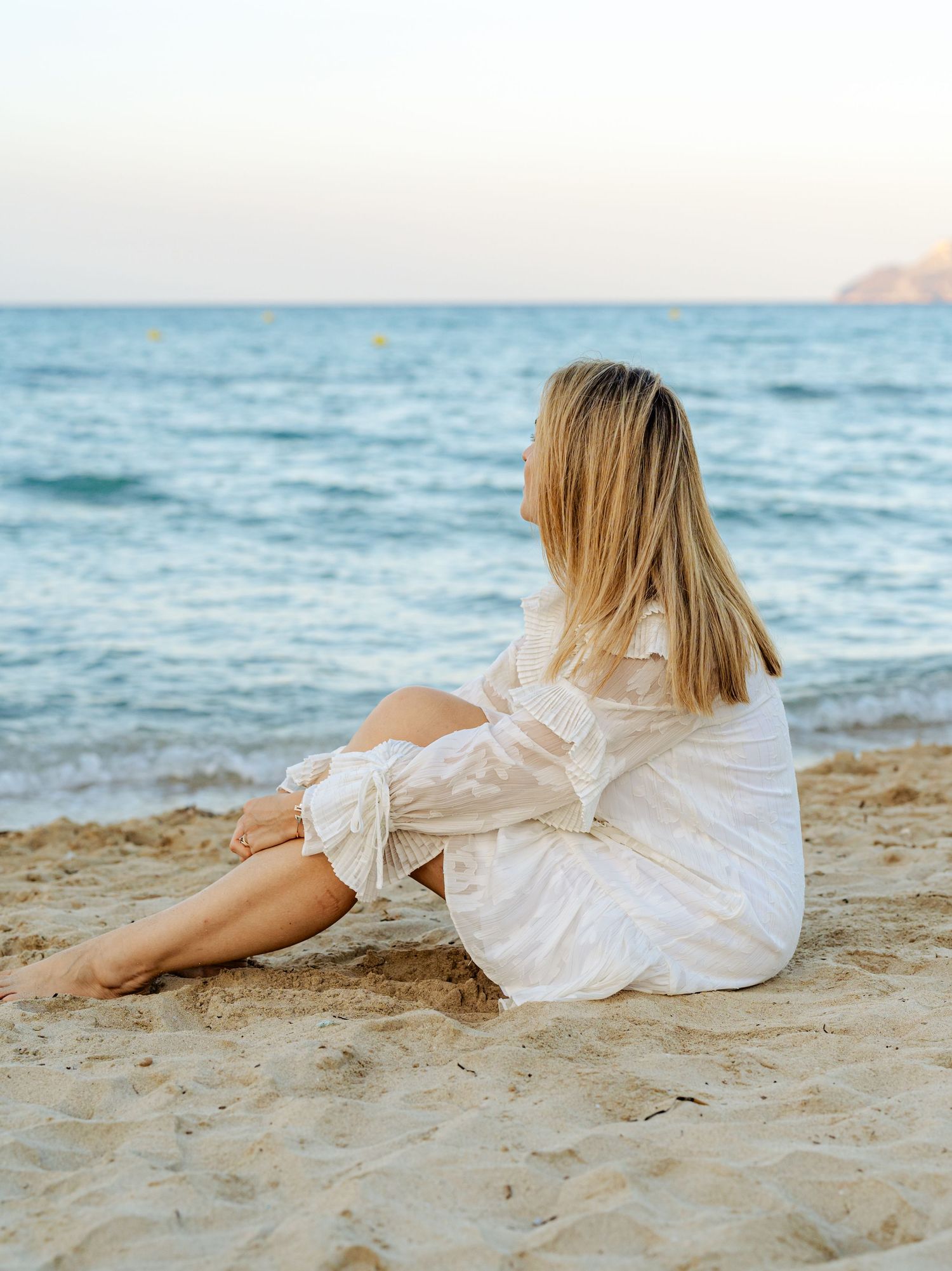 Young blonde woman in a white dress sitting on sandy beach gazing at the calm blue ocean at sunset.