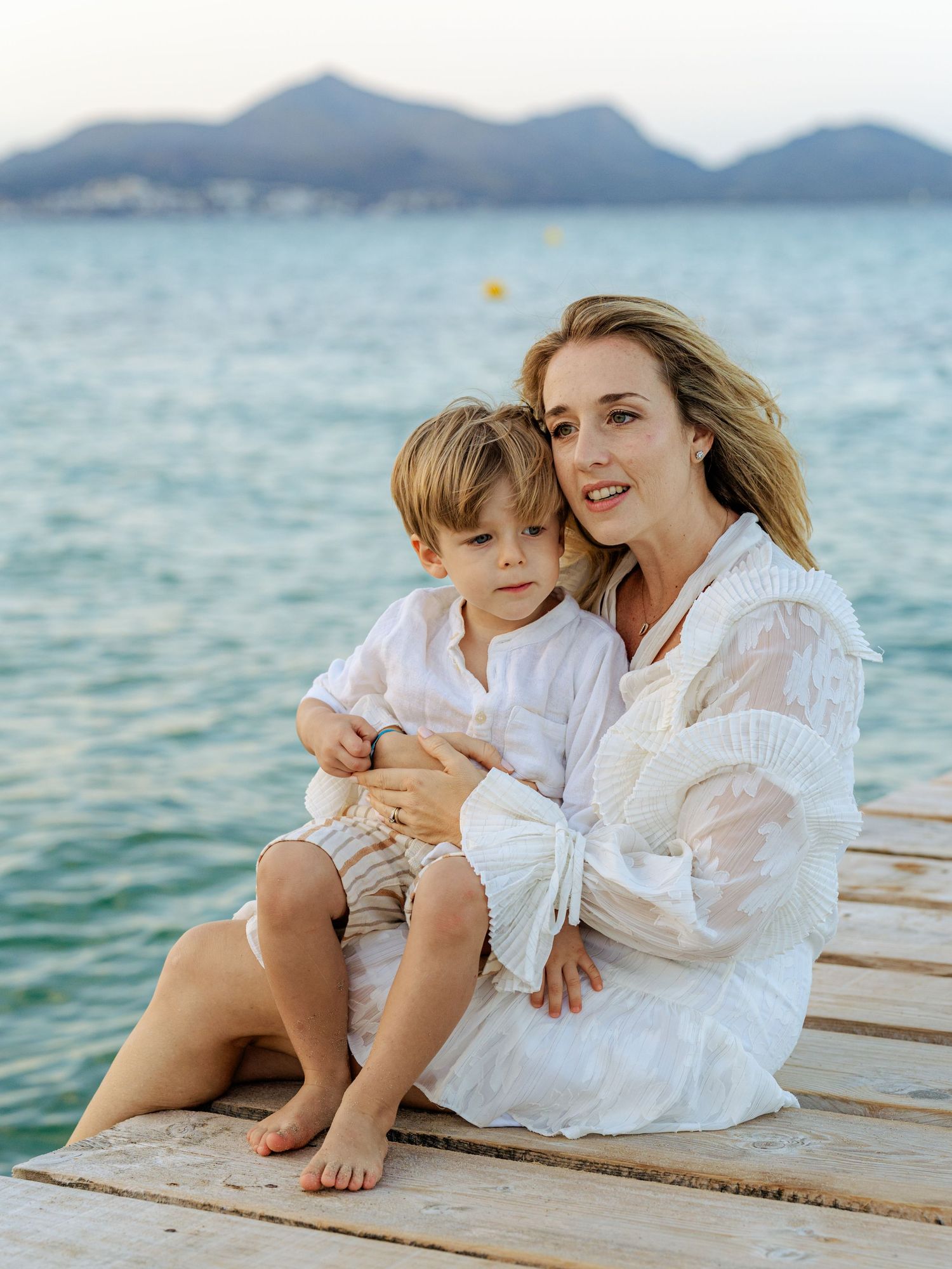 Loving mother sitting barefoot on a wooden pier hugging her young son with ocean backdrop.