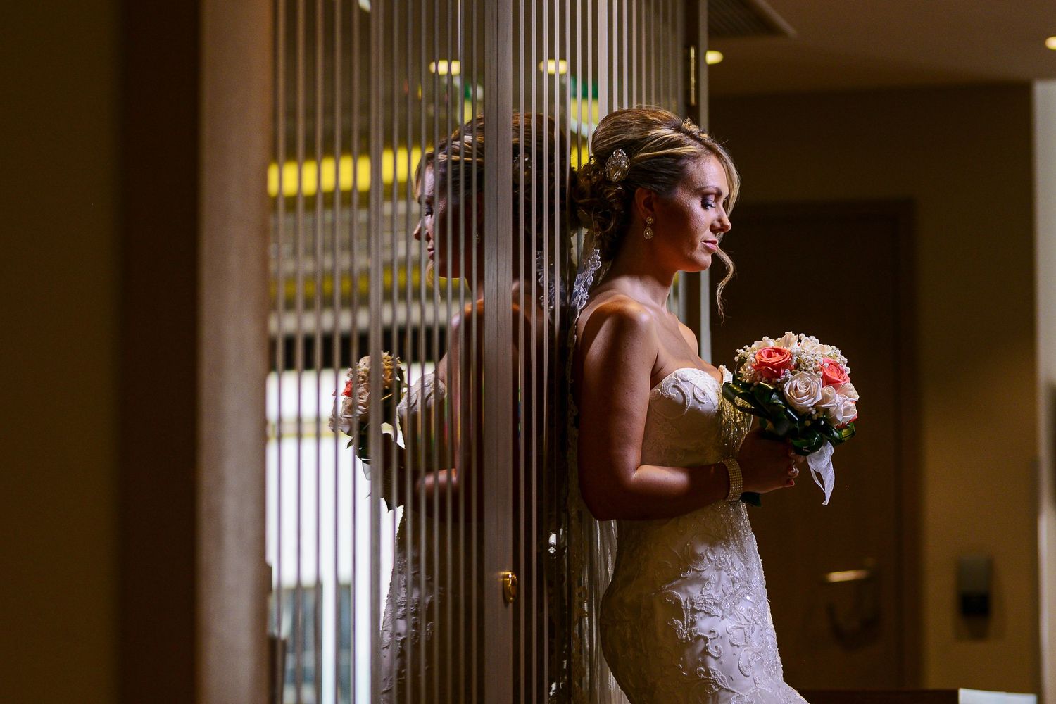 Bride in lace wedding dress holding a bouquet, reflected in decorative metal panels indoors.