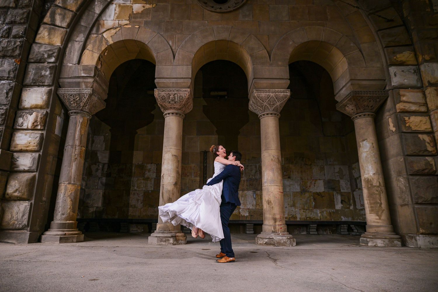 Groom lifting bride in white gown under stone arches of historic building during wedding photo session.