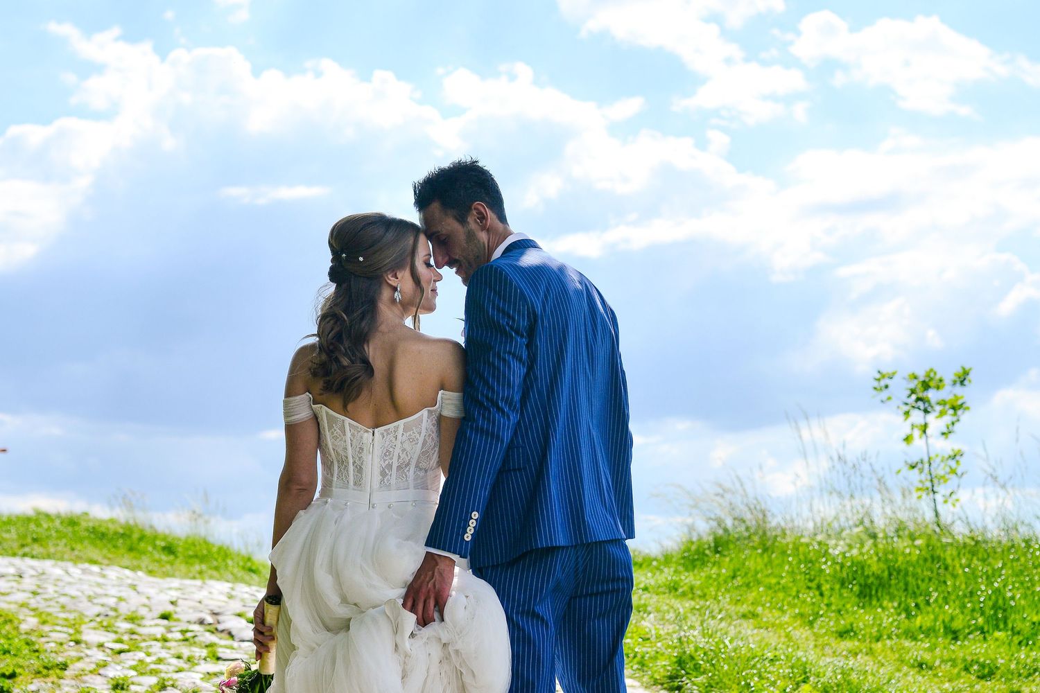 Bride and groom sharing a tender moment outdoors with lush green hills and blue sky behind them.