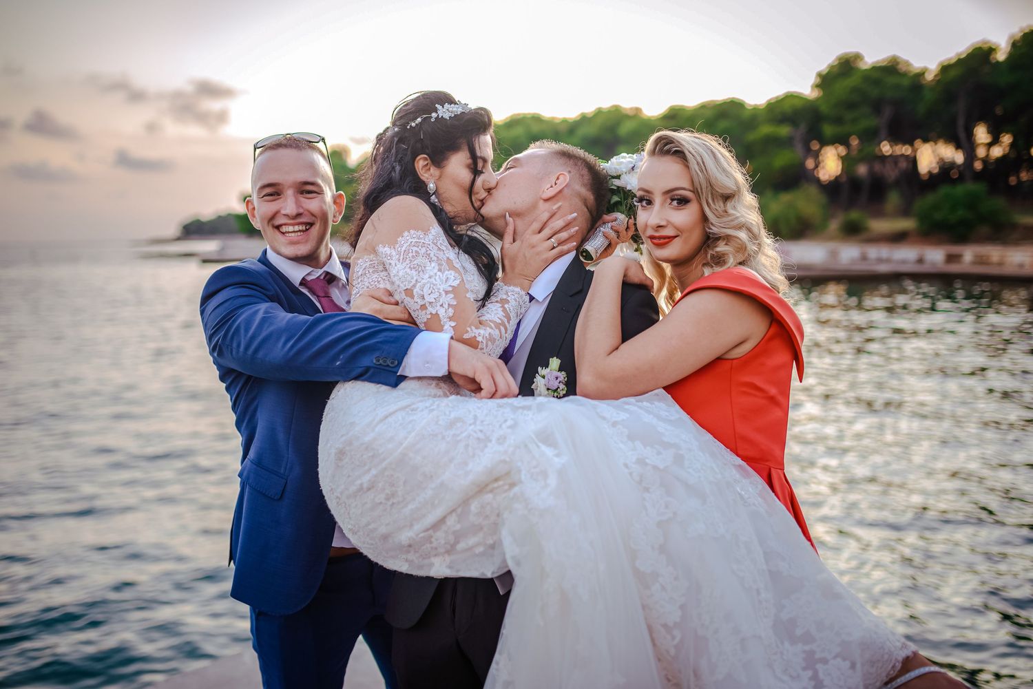Joyful wedding party photo with bride and groom being lifted by guests near sparkling waterfront at sunset.