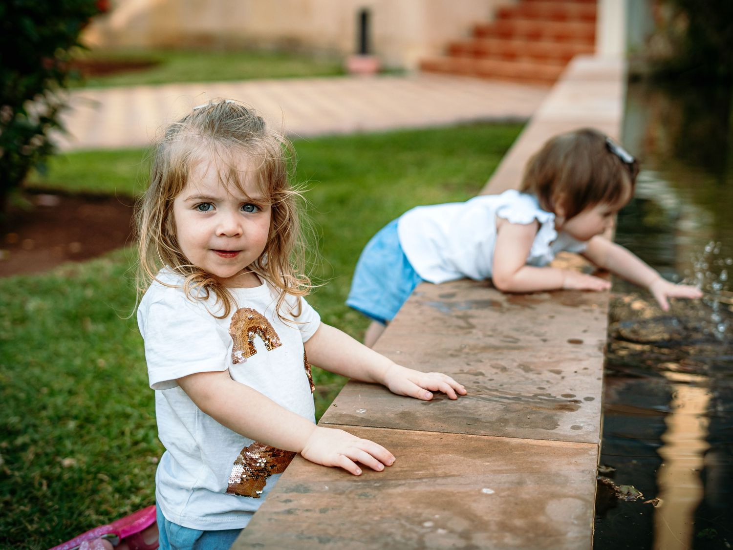 Two little girls exploring a garden water feature, older girl smiling at camera.