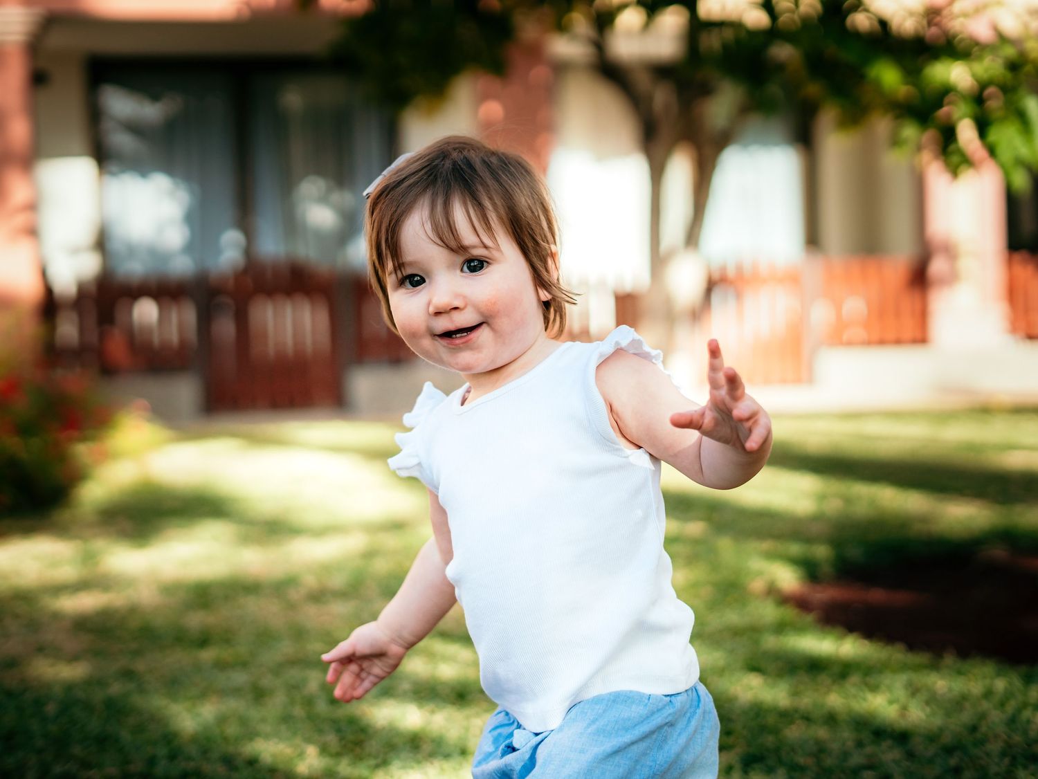 Cheerful toddler girl in white top and blue shorts reaching out while playing on green grass.