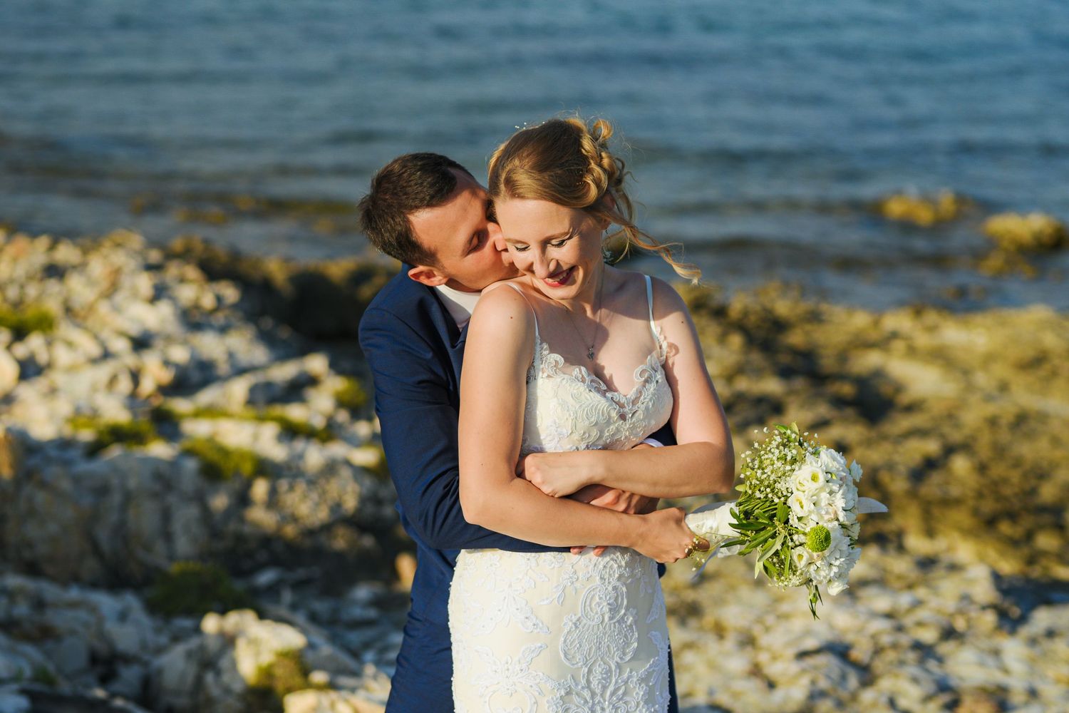 Groom tenderly kisses bride on the cheek as she smiles holding a white bouquet by a rocky shoreline.