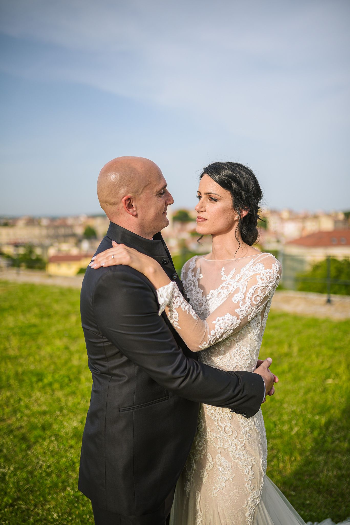Bride in lace gown and groom in black suit embrace face to face on a green hillside.