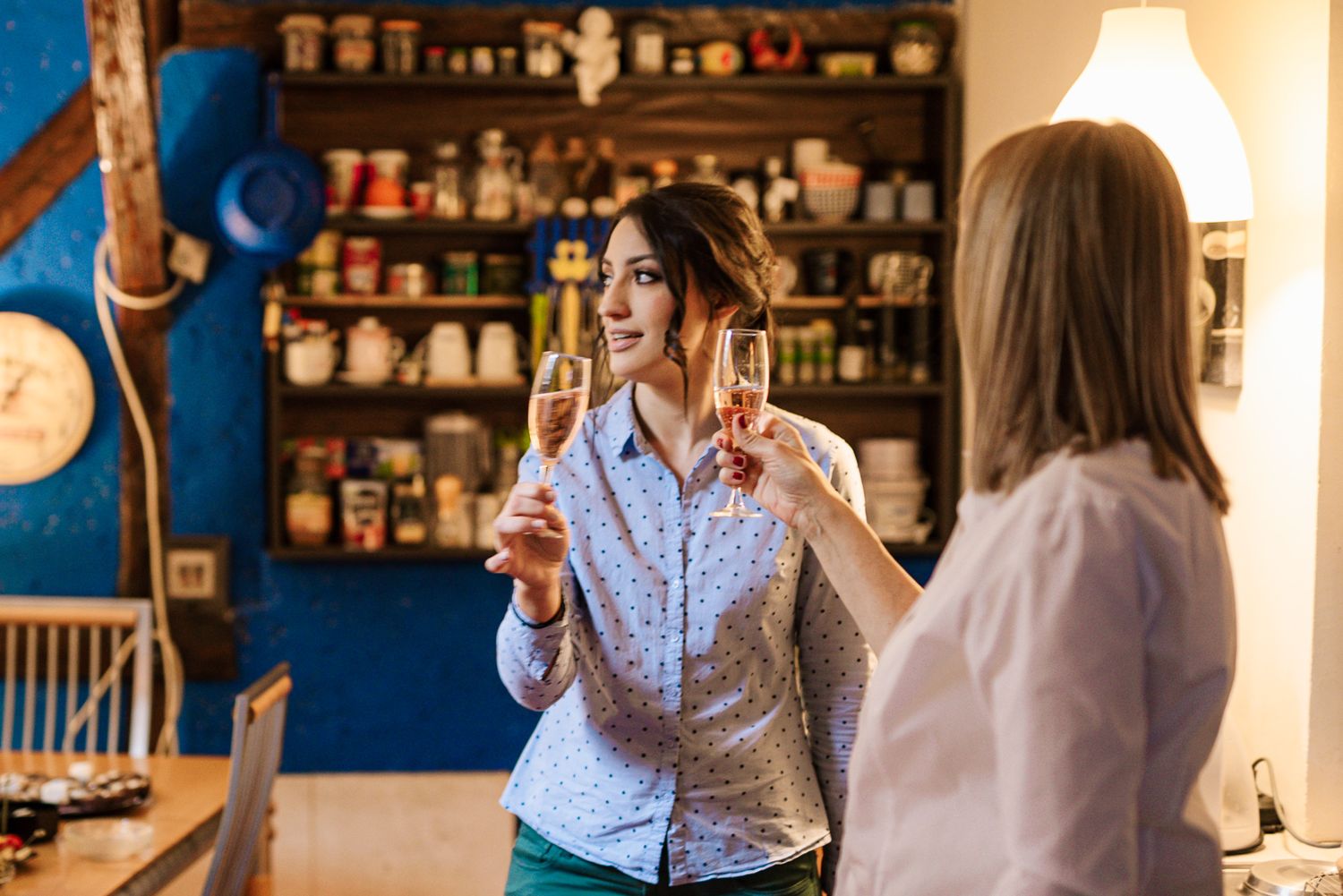 Two women celebrating with champagne glasses in a cozy kitchen decorated with colorful shelves.