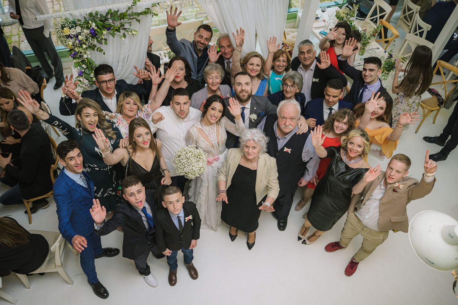 Large wedding party group photo with bride and groom surrounded by smiling family and friends.