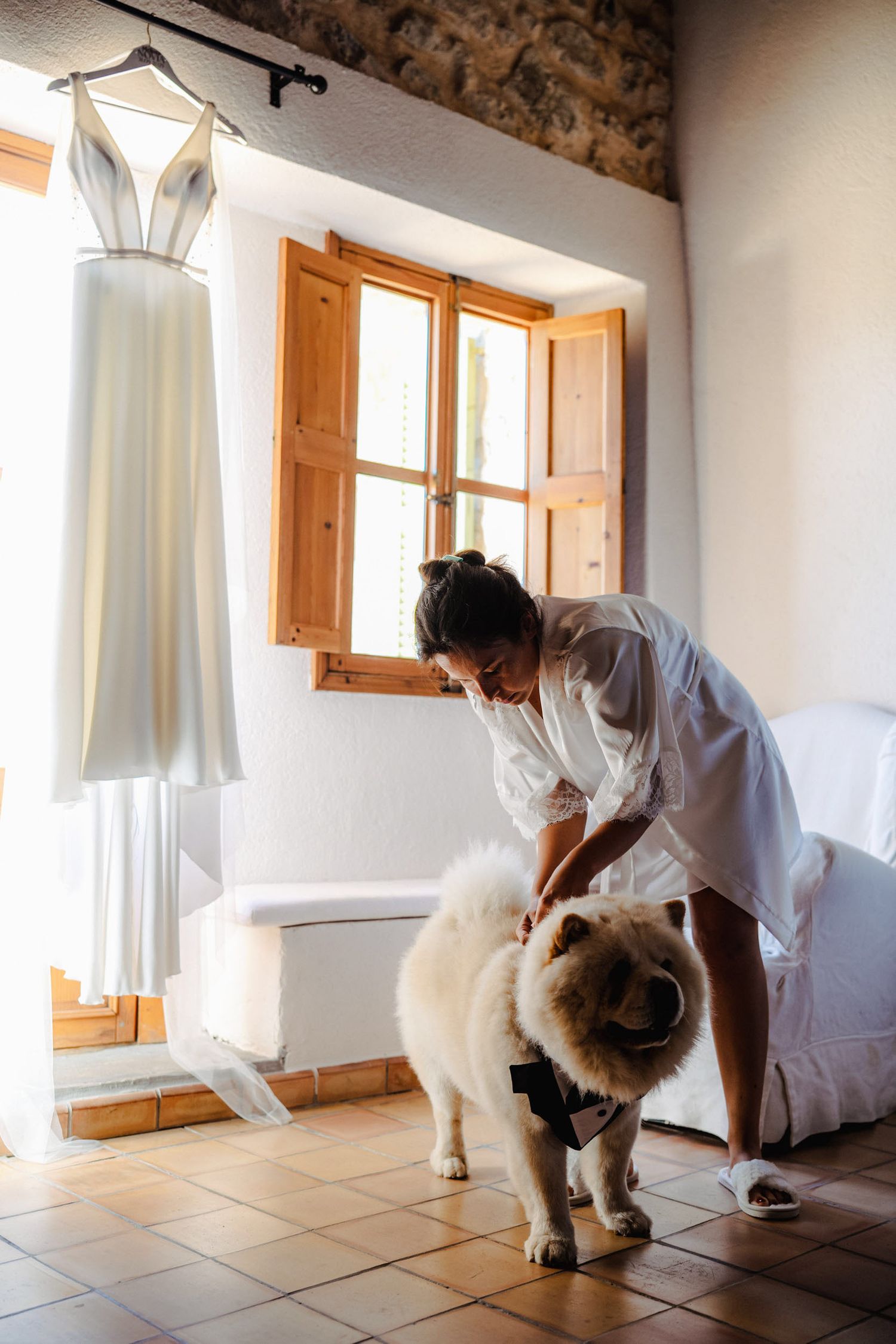 Bride in white robe petting fluffy white dog beside hanging wedding dress near sunlit window.
