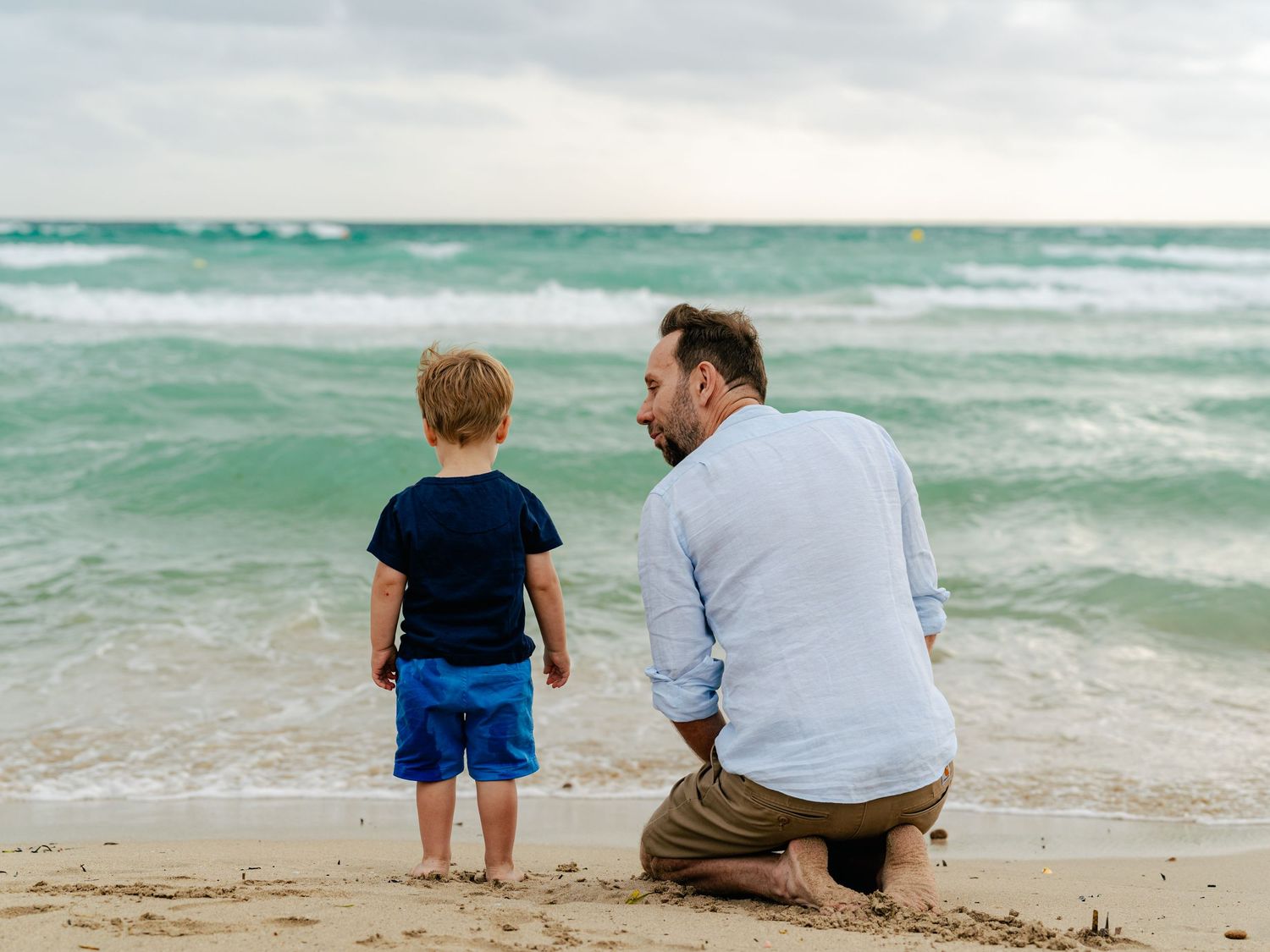 Father and toddler son sitting together on sandy beach watching ocean waves.