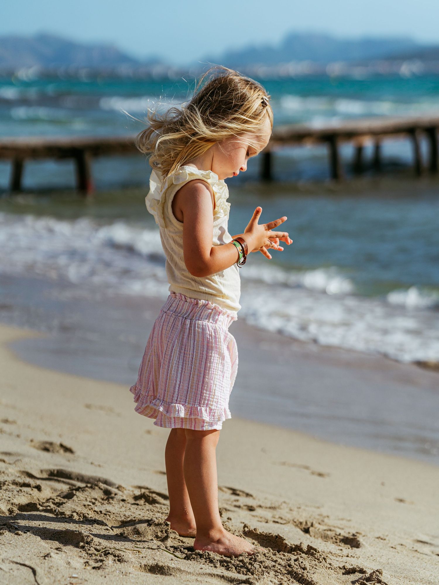 Little blonde girl in a pink skirt standing barefoot on a sunny beach examining her hands.