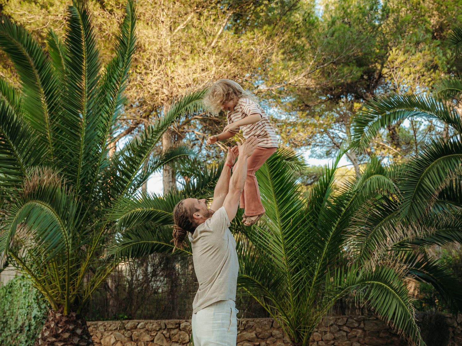 Dad joyfully lifting daughter above his head among green palm trees in sunny garden.