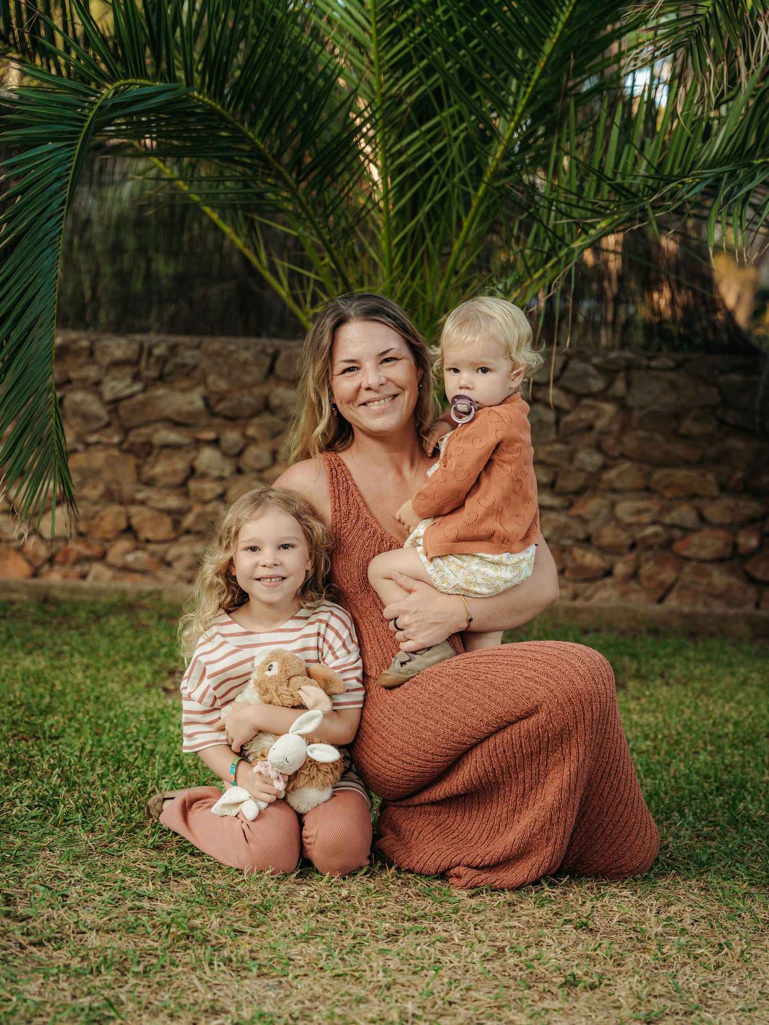 Smiling mother in rust dress posing with two young children and a bunny outdoors.