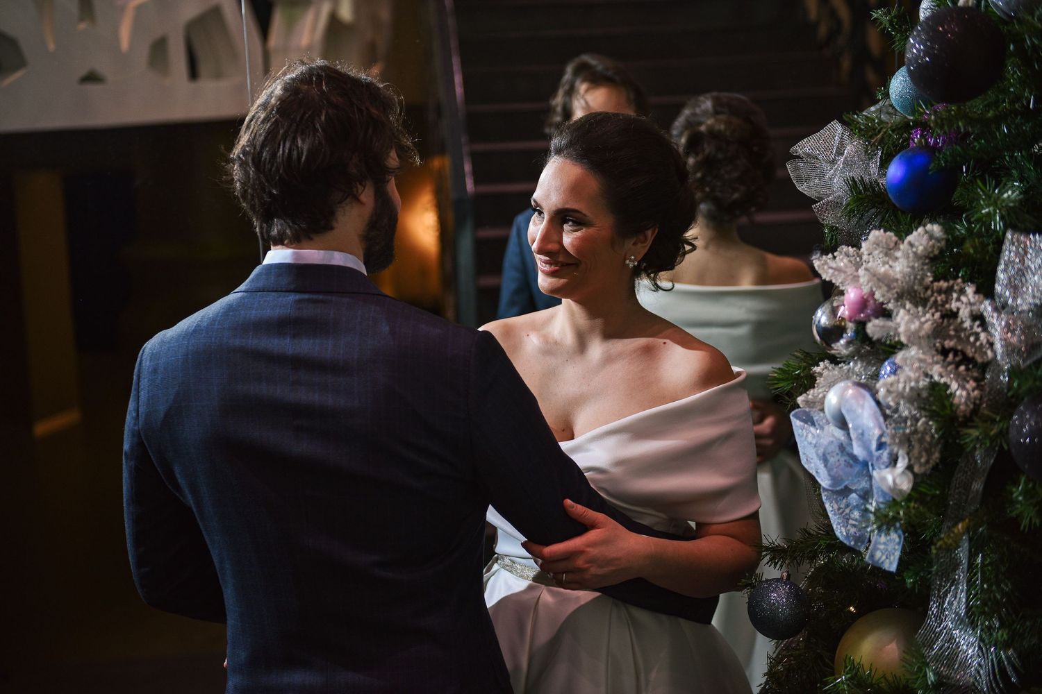 Couple shares an intimate moment near a decorated Christmas tree at their elegant winter wedding ceremony.