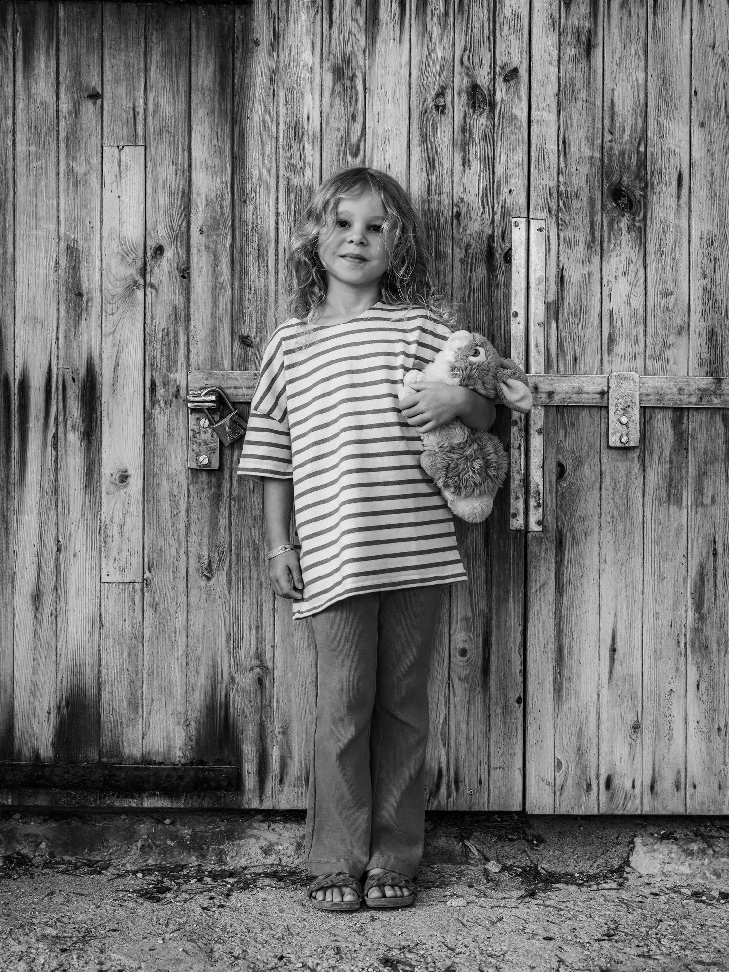 Black and white portrait of young girl with stuffed bear standing before aged wooden barn doors.