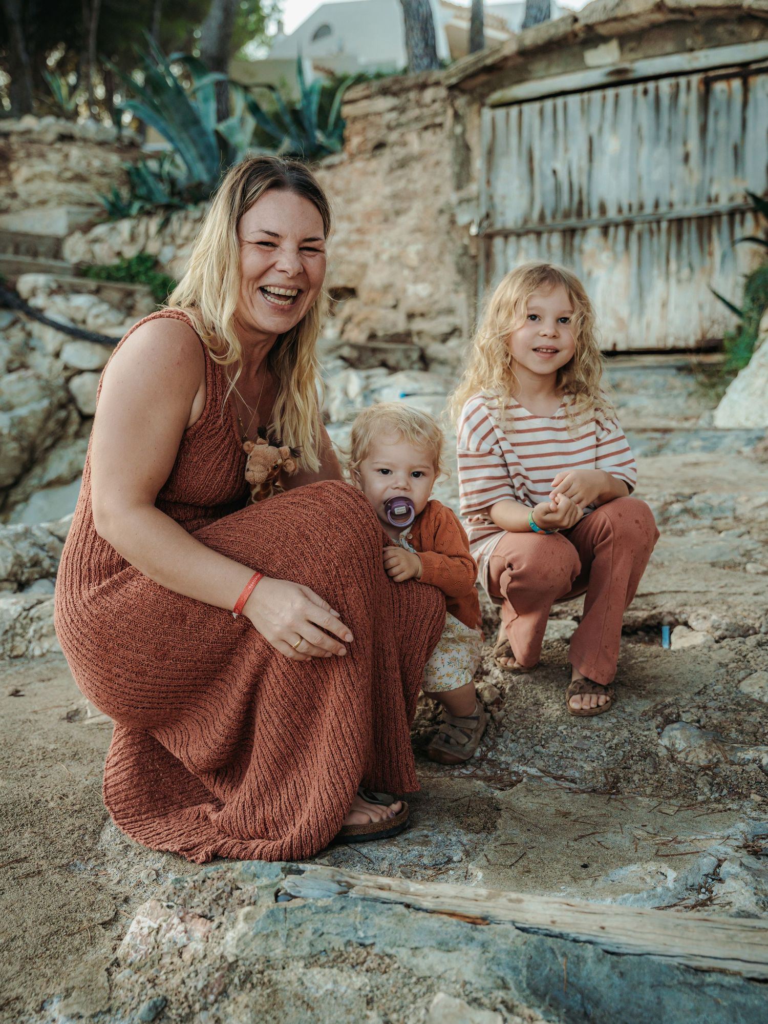 A laughing blonde woman in rust dress poses with two young children on rocky ground outdoors.