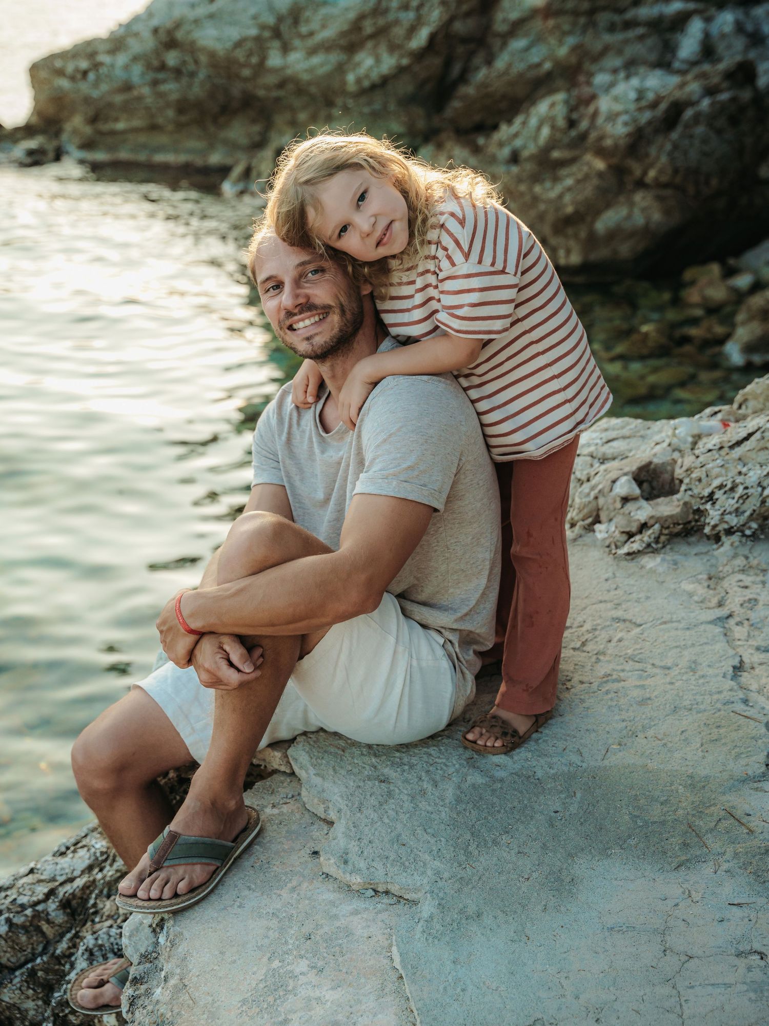 A smiling father sits on rocks by the water as his young daughter leans over his shoulder affectionately.