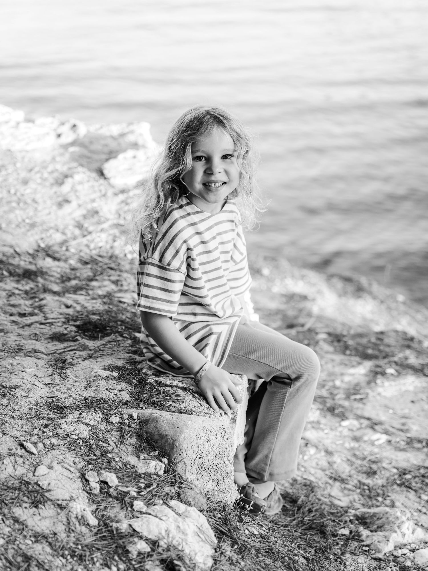 Black and white portrait of a blonde girl sitting on rocks by the sea.