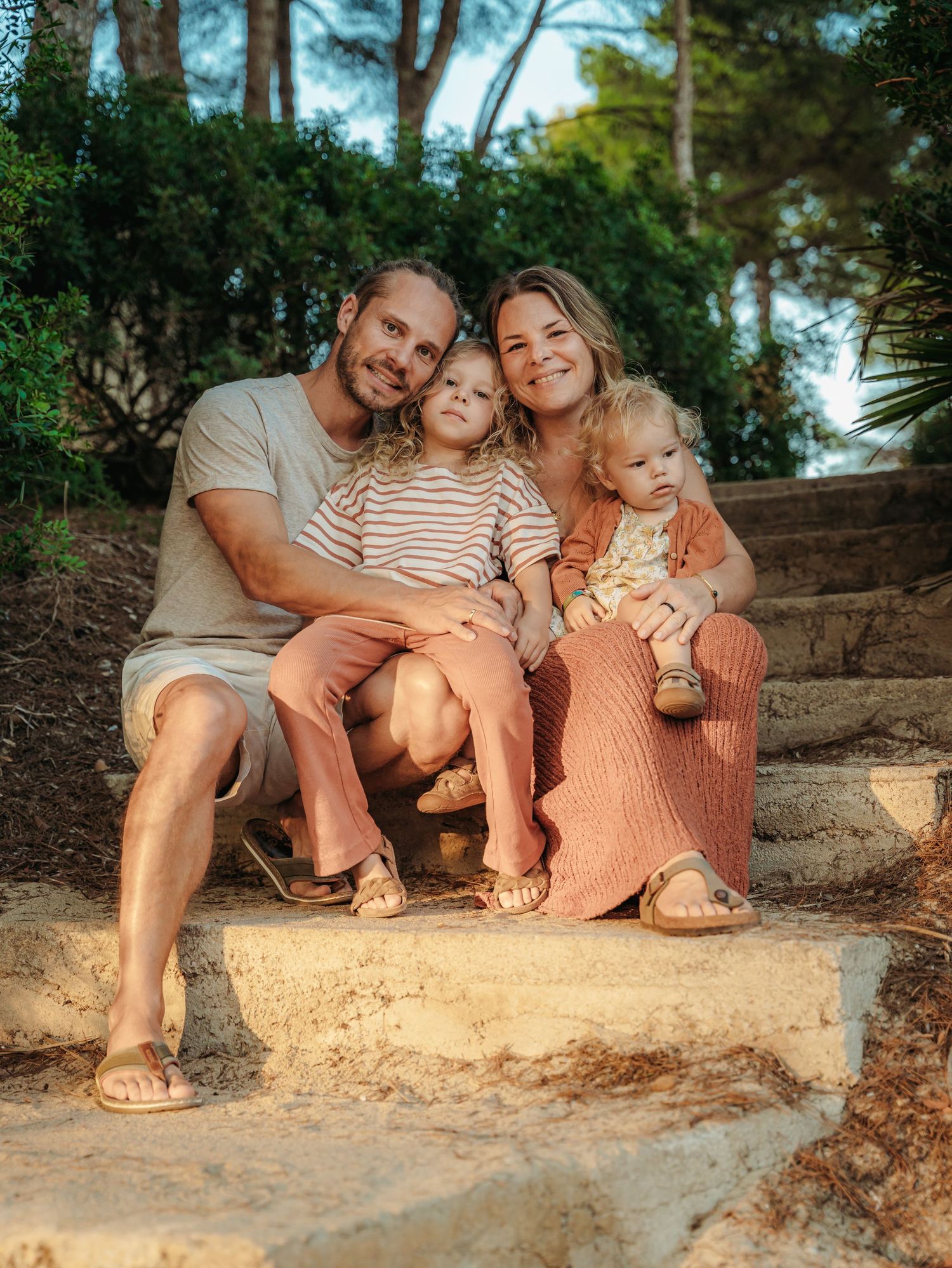 Happy family of four sitting on stone steps surrounded by lush greenery during golden hour.