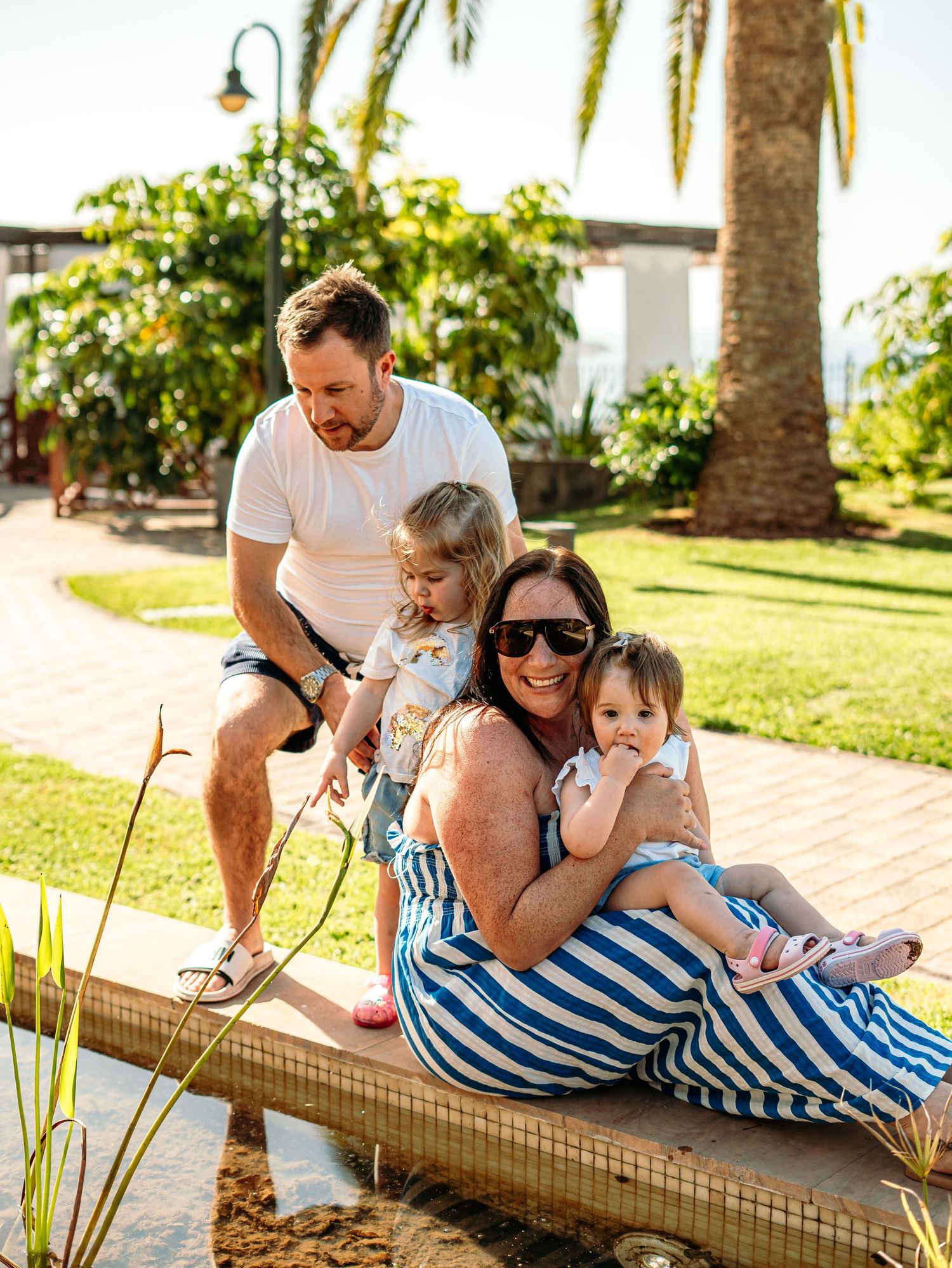 Candid family moment with parents and two children relaxing by a sunny garden pond.