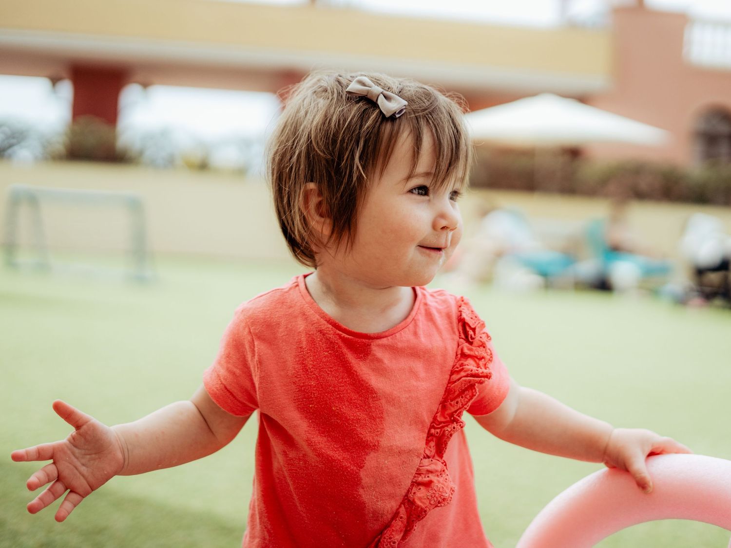 Cheerful toddler girl in red ruffled top with arms outstretched playing on green grass.