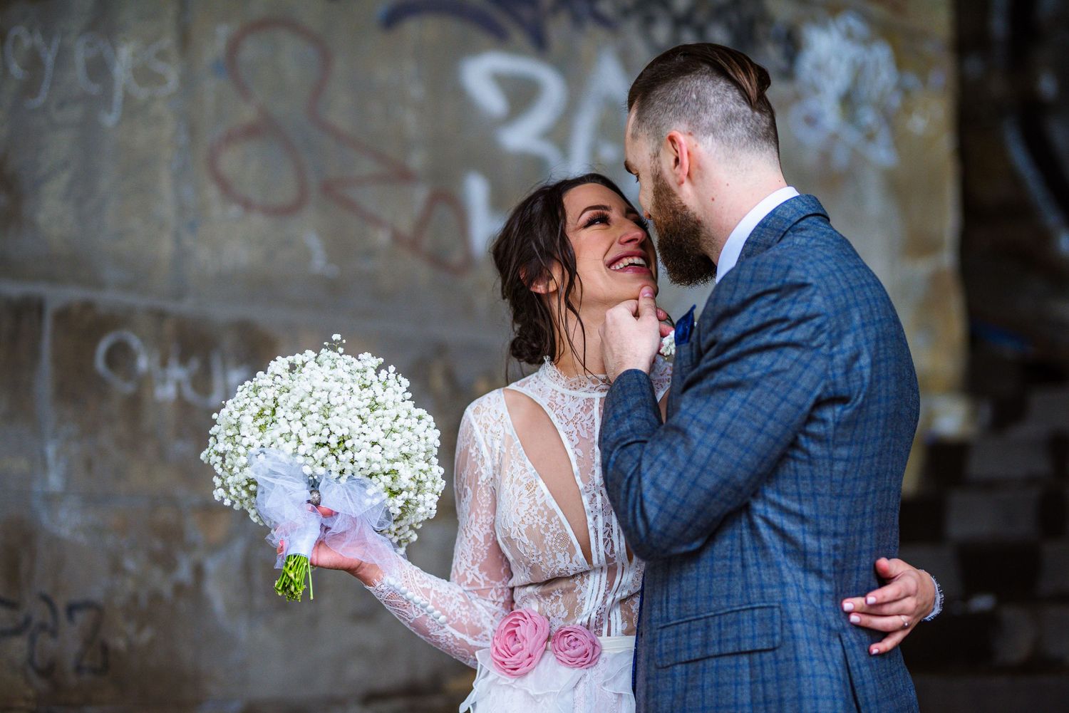 Smiling bride holding white bouquet gazes at bearded groom in blue plaid suit against graffiti wall.