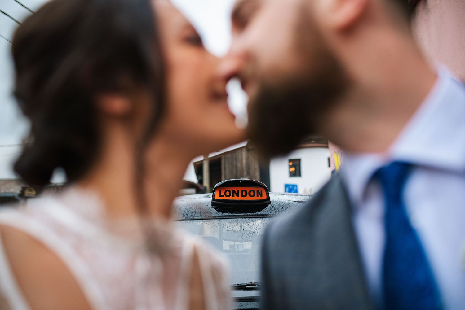 Couple sharing romantic kiss behind classic London taxi cab sign in soft focus wedding portrait.