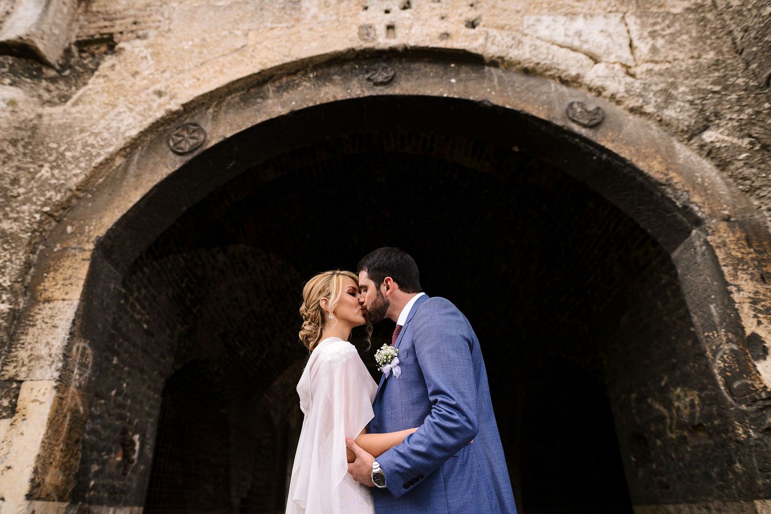 Bride and groom kissing beneath an ancient stone arch - love framed by history and time. Herzenshaus Studio, Frankfurt.