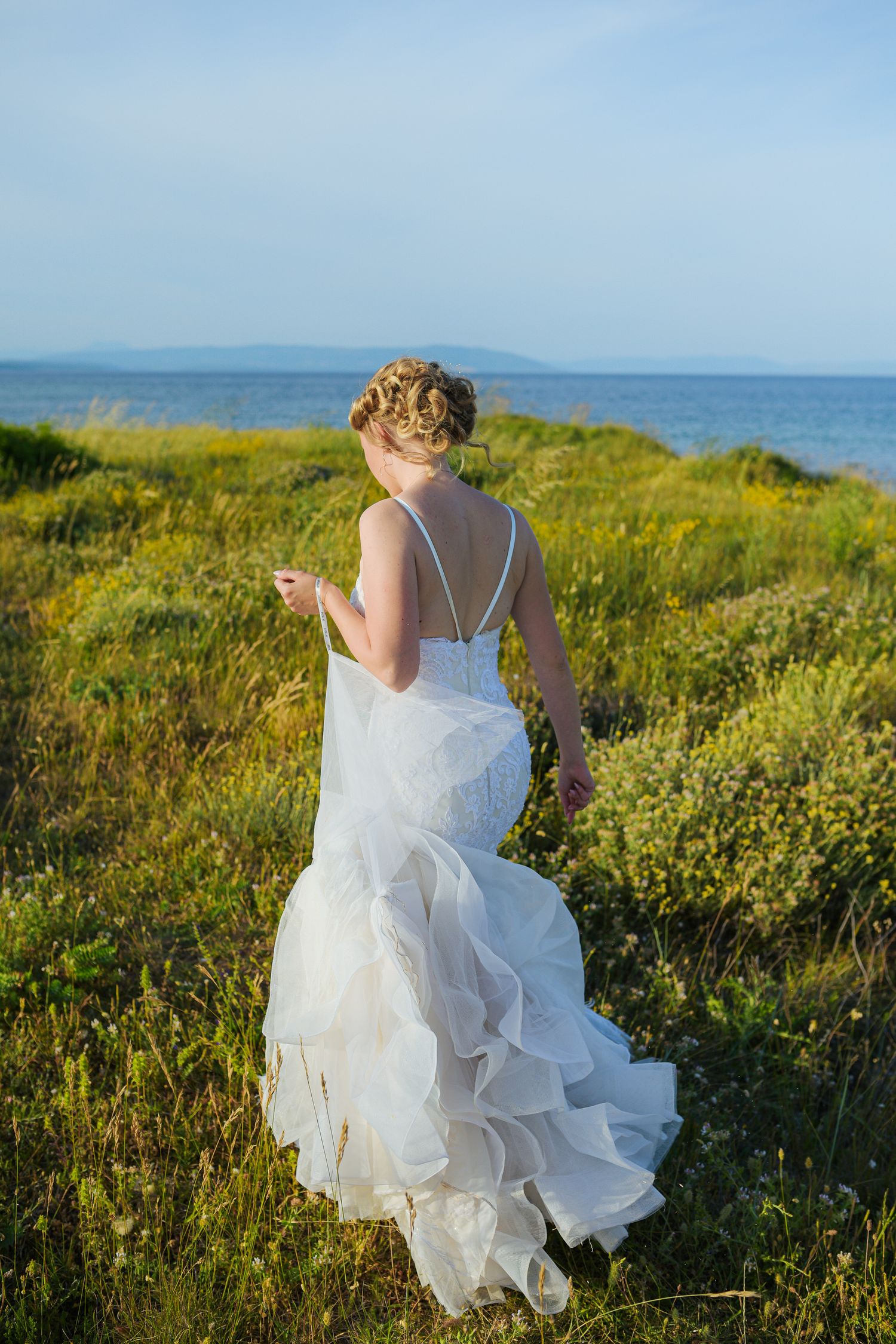 Elegant bride in a ruffled white gown walks through a coastal meadow with the blue sea in the distance.