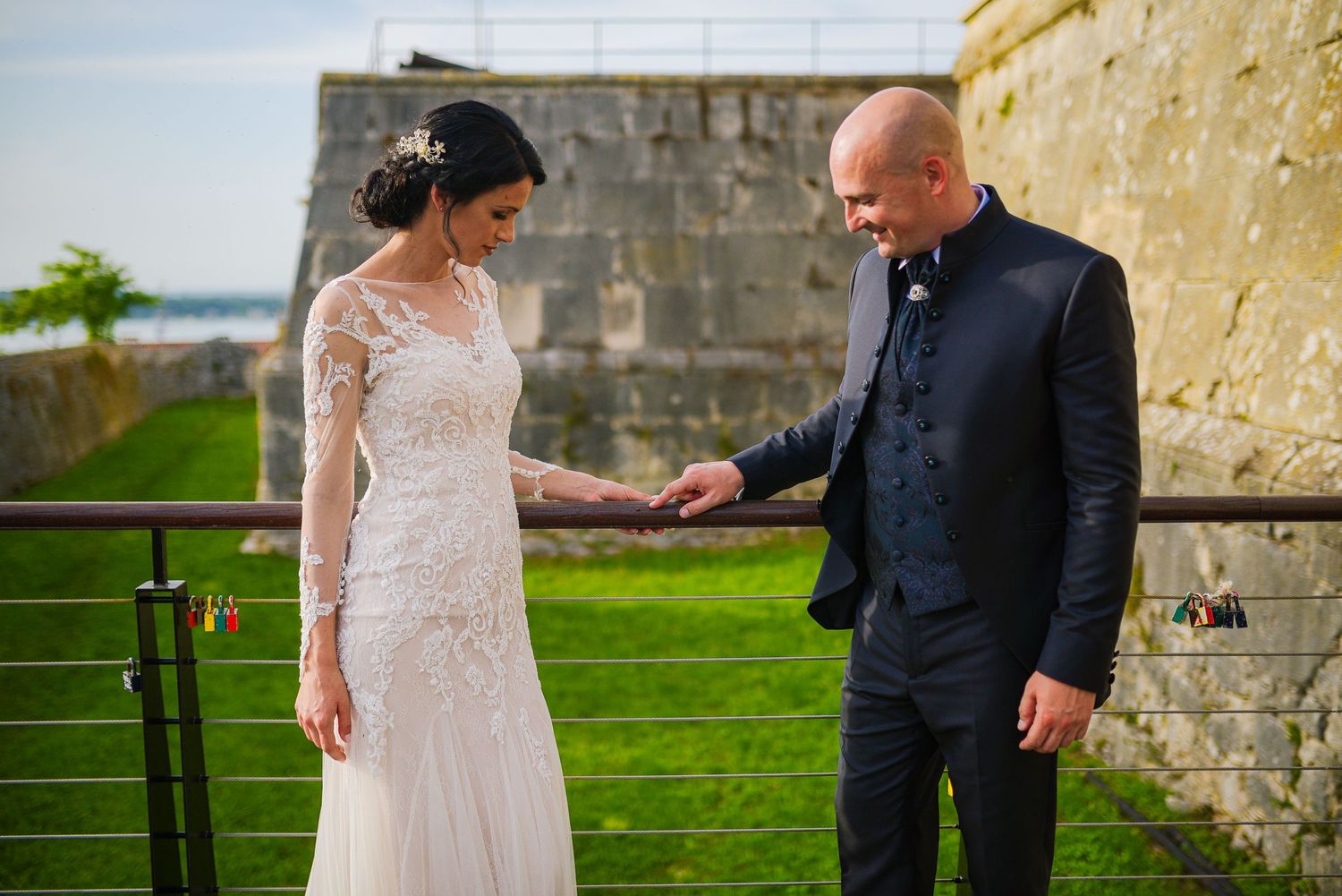 Bride in lace gown and groom in navy suit hold hands on a railing at an outdoor wedding venue.