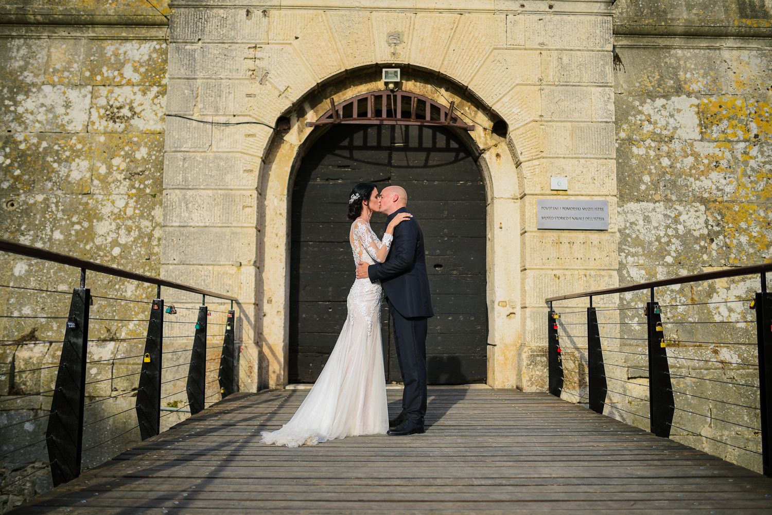 Newlyweds sharing a romantic kiss on a wooden bridge before a historic stone gate.